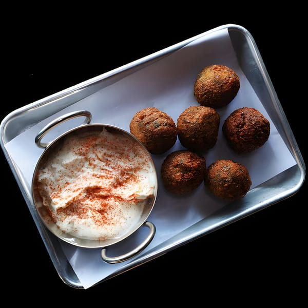 A metal tray with six Twisted Greek falafel balls and a bowl of tzatziki sauce topped with paprika.