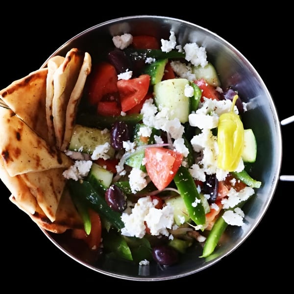 A vibrant Twisted Greek salad with feta, olives, tomatoes, cucumbers, and pita bread in a metal bowl.