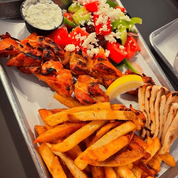 Grilled chicken skewers, fries, Greek salad with feta, pita bread, and tzatziki sauce on a tray.